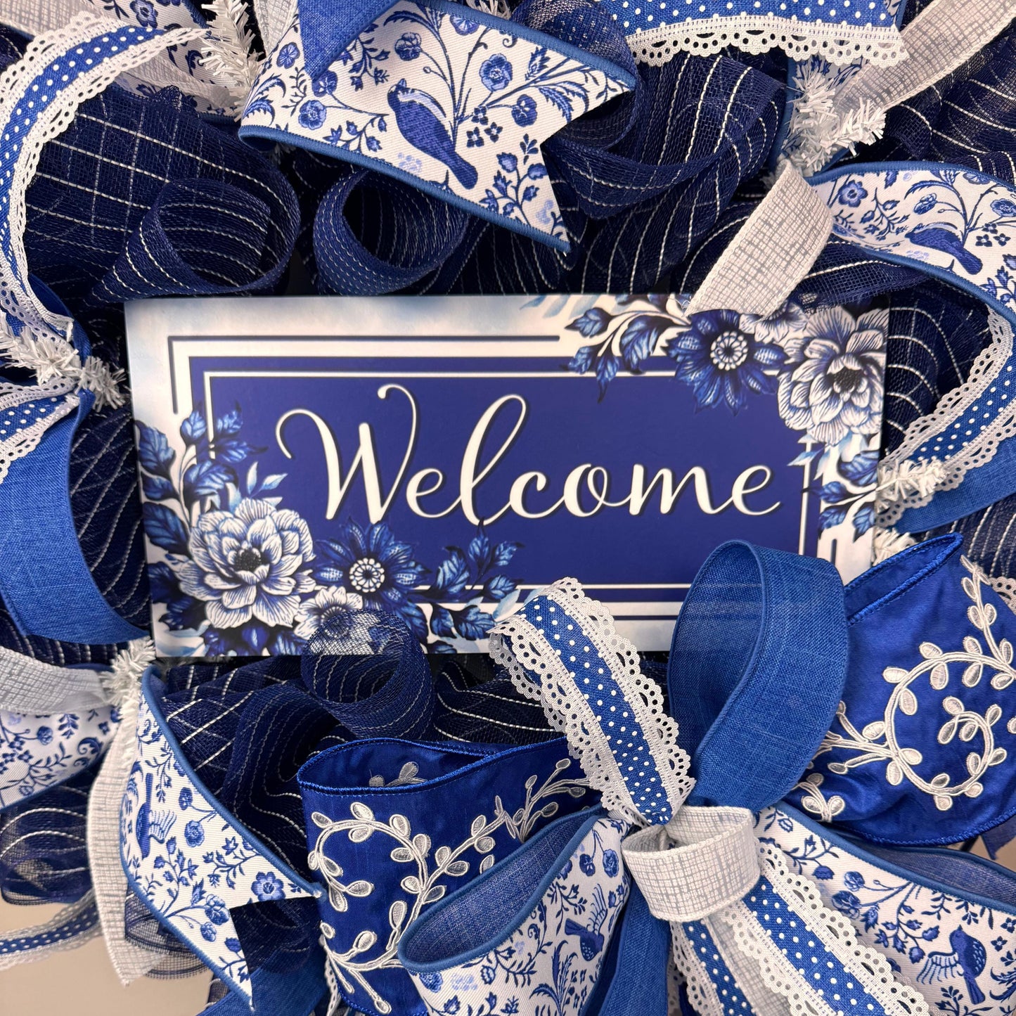 Close-up of blue welcome sign with floral detail on a handmade ribbon wreath, featuring intricate chinoiserie-inspired design elements.