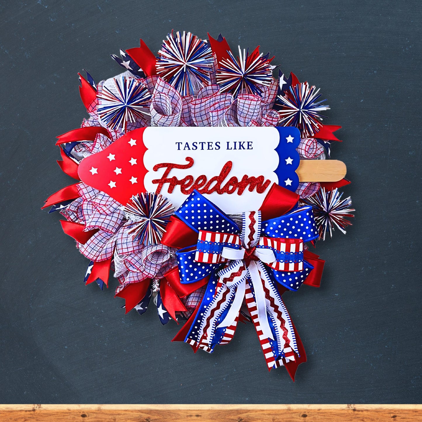 Handmade patriotic door wreath on navy blue background, perfect for classroom, porch, or home entryway, with red white and blue bows and Tastes Like Freedom sign