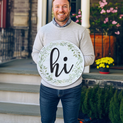 Man holding 18-inch round wooden hi door sign with greenery wreath on front porch, perfect size demonstration for entryway decor