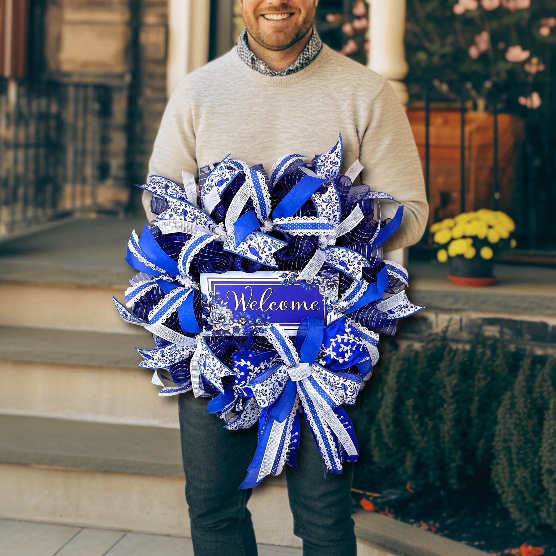 Man holding a handmade blue and white welcome wreath, showing scale and lush designer ribbon for year-round entryway decor.