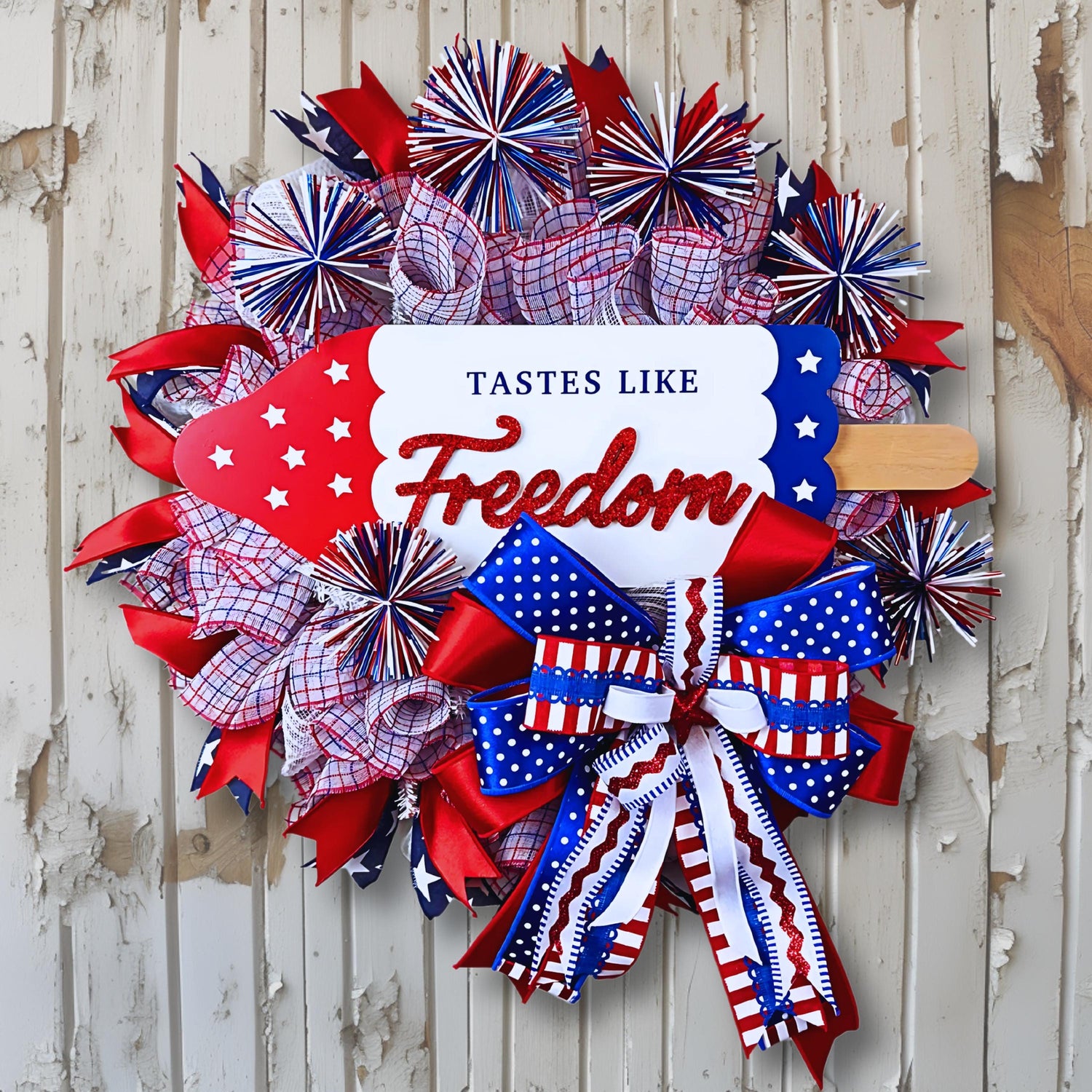 Patriotic red white and blue front door wreath for 4th of July with rustic wood background, handmade with bows and glitter sign that says Tastes Like Freedom